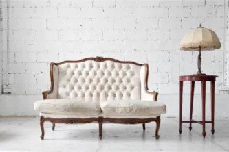 Ghế sofa cổ điển bọc nệm trắng với khung gỗ chạm khắc tinh xảo, đặt cạnh bàn tròn cao và đèn kim loại chao kem có tua rua.